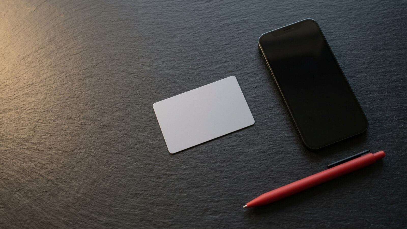 A blank plastic card, a red pen and an iPhone with a dark screen on a dark slate desk
