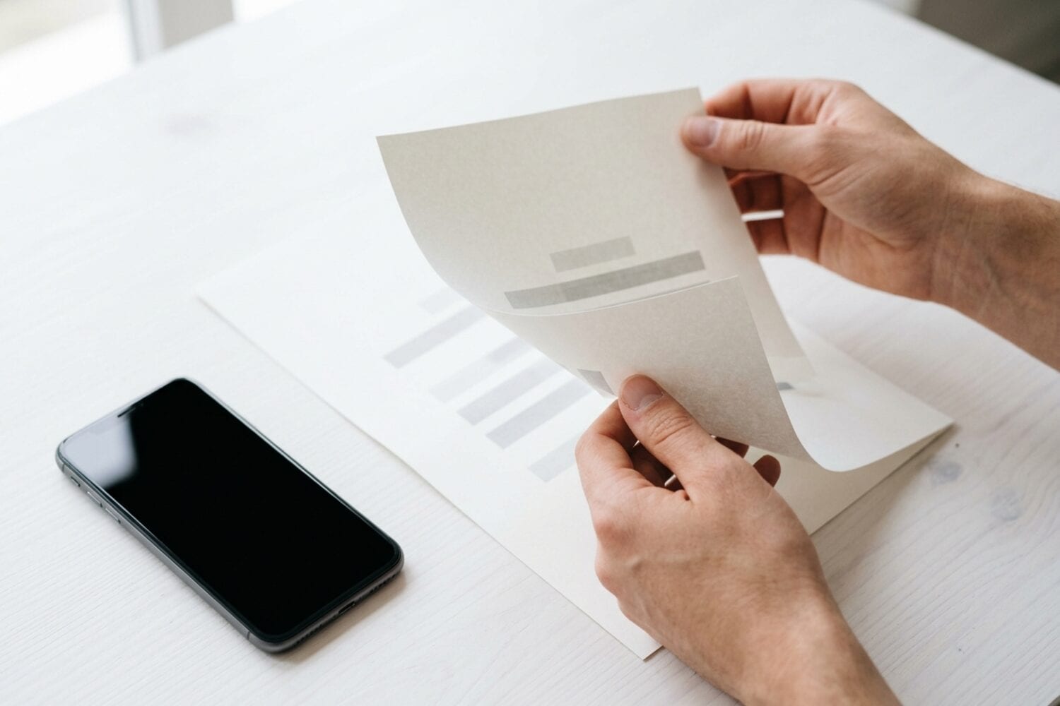 Dos manos dan la vuelta a una hoja impresa sobre un escritorio blanco, con un iPhone de pantalla oscura al lado
