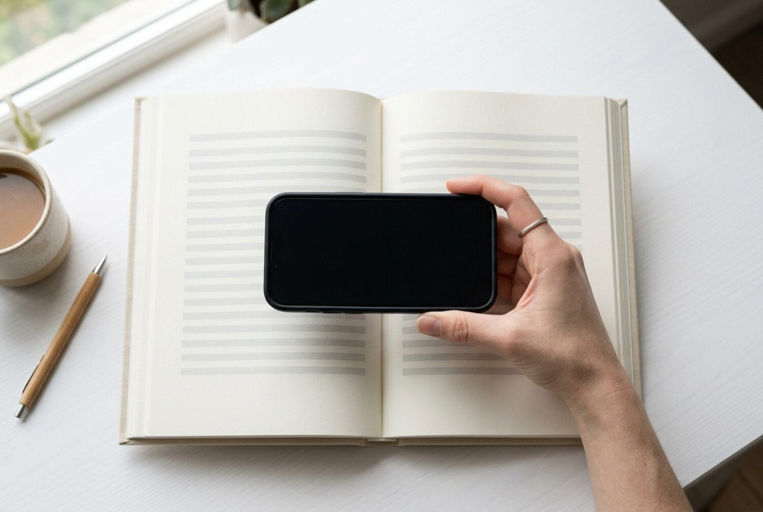 Eine Hand hält ein iPhone flach über ein aufgeschlagenes gebundenes Buch auf einem weißen Schreibtisch, um eine Doppelseite zu scannen