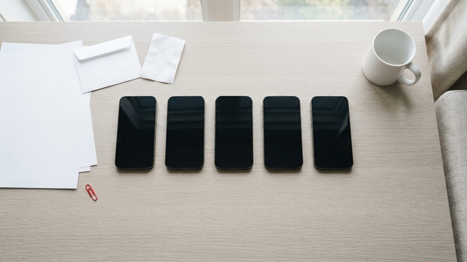 Cinq iPhone aux écrans sombres alignés sur un bureau clair avec du papier vierge, un ticket de caisse et une enveloppe